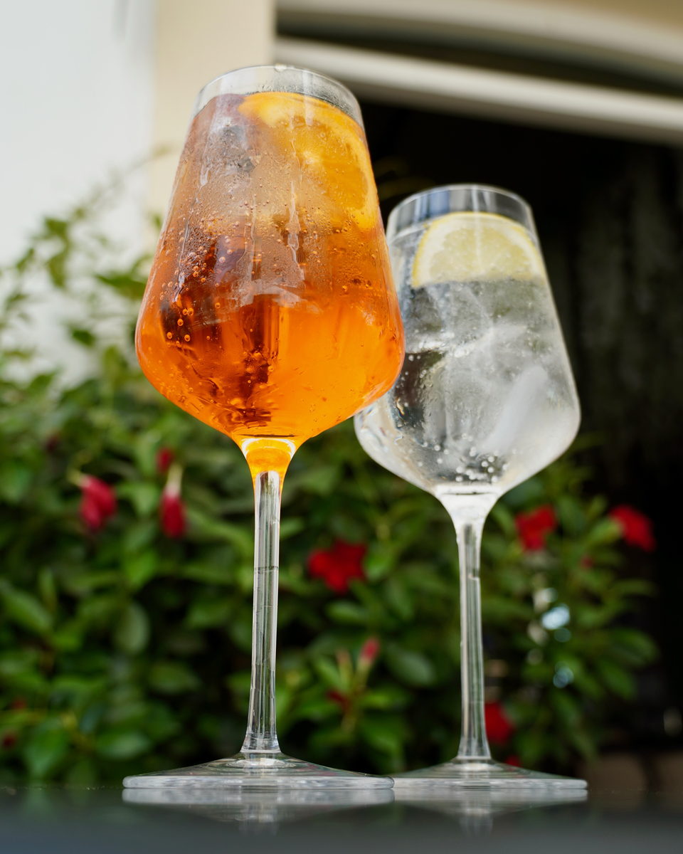 A spritz and a gin and tonic, beaded with condensation, on a table in front of flowering shrubs.