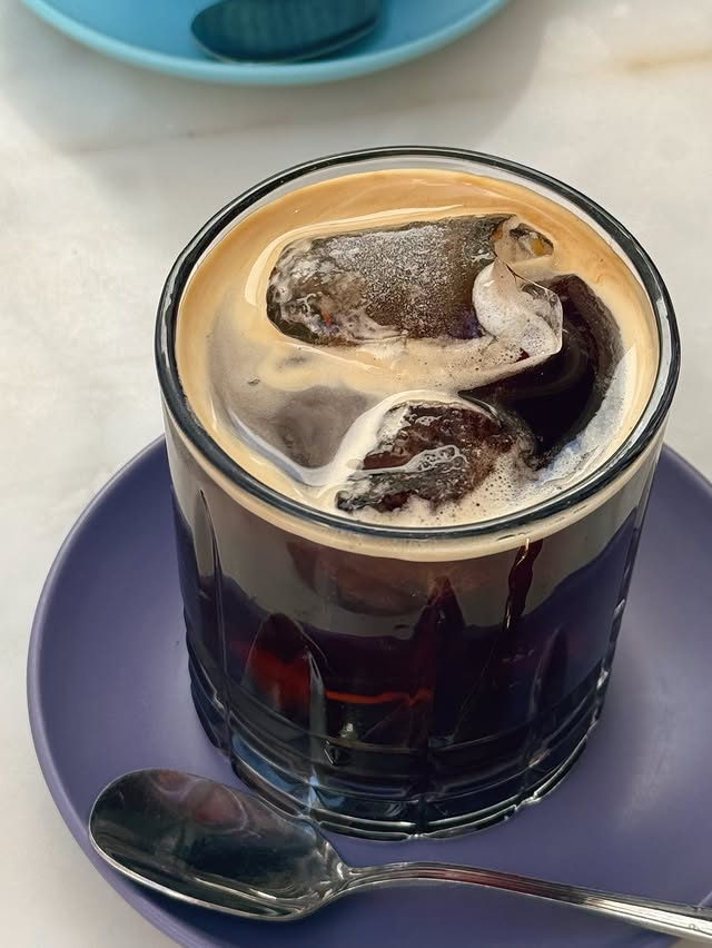 Iced coffee in a heavy tumbler with crema on top, on a lilac saucer.