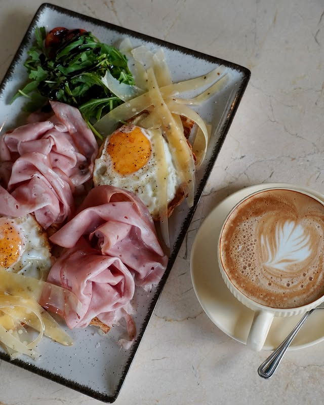 Ruffled mortadella and fried eggs with shaved cheese and rocket on a dark speckled plate, beside a cappuccino.