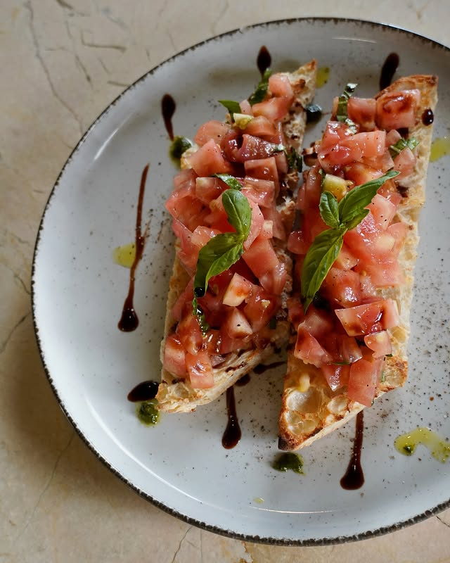 Two slices of bruschetta piled with chopped tomato and basil, dressed with balsamic and pesto.