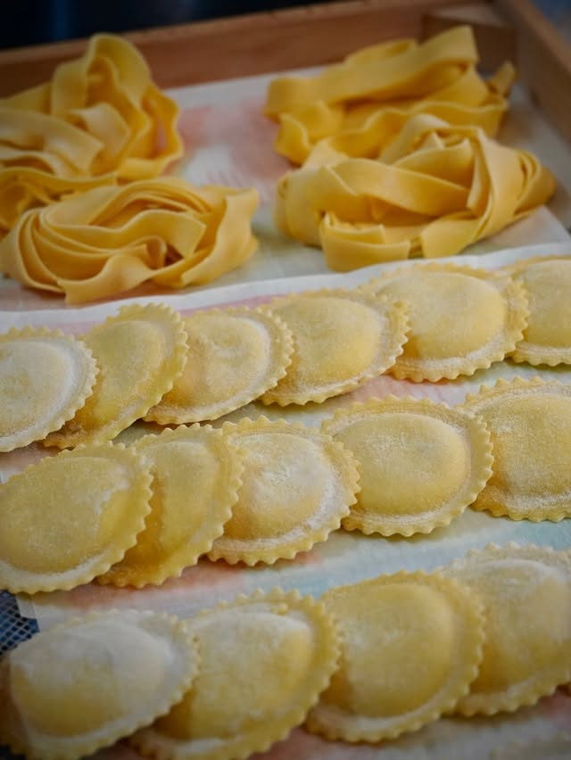 Nests of fresh pappardelle and rows of ravioli laid out on a wooden tray.