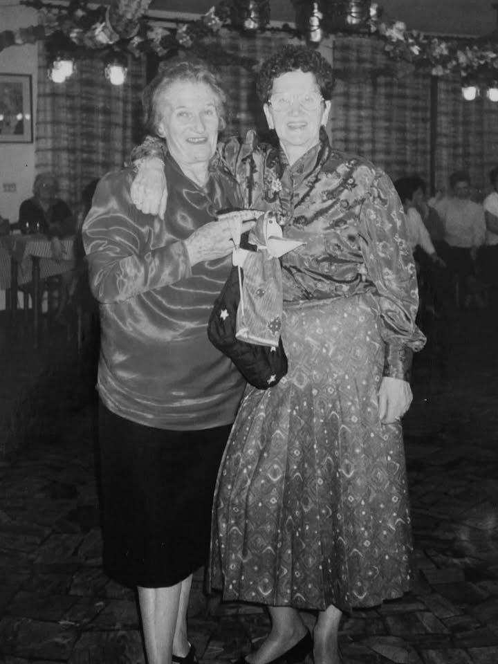 Two women arm in arm on the floor of a dance hall, dressed up for the evening.