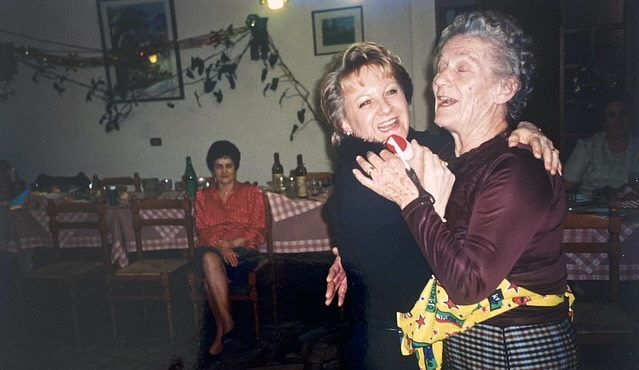 An older woman in an apron dancing cheek to cheek with a younger woman.