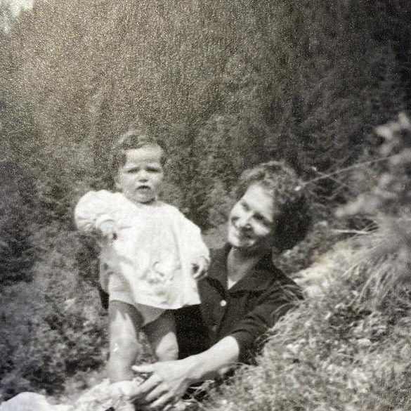 A woman sitting on a hillside, steadying a small child on its feet beside her.