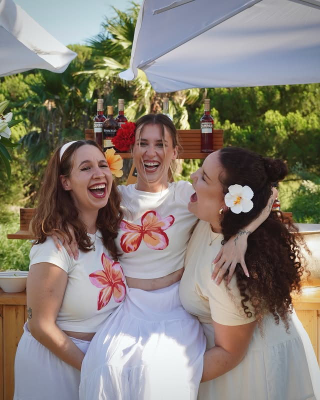 Three friends in white and cream, flowers in their hair, laughing together under a parasol.