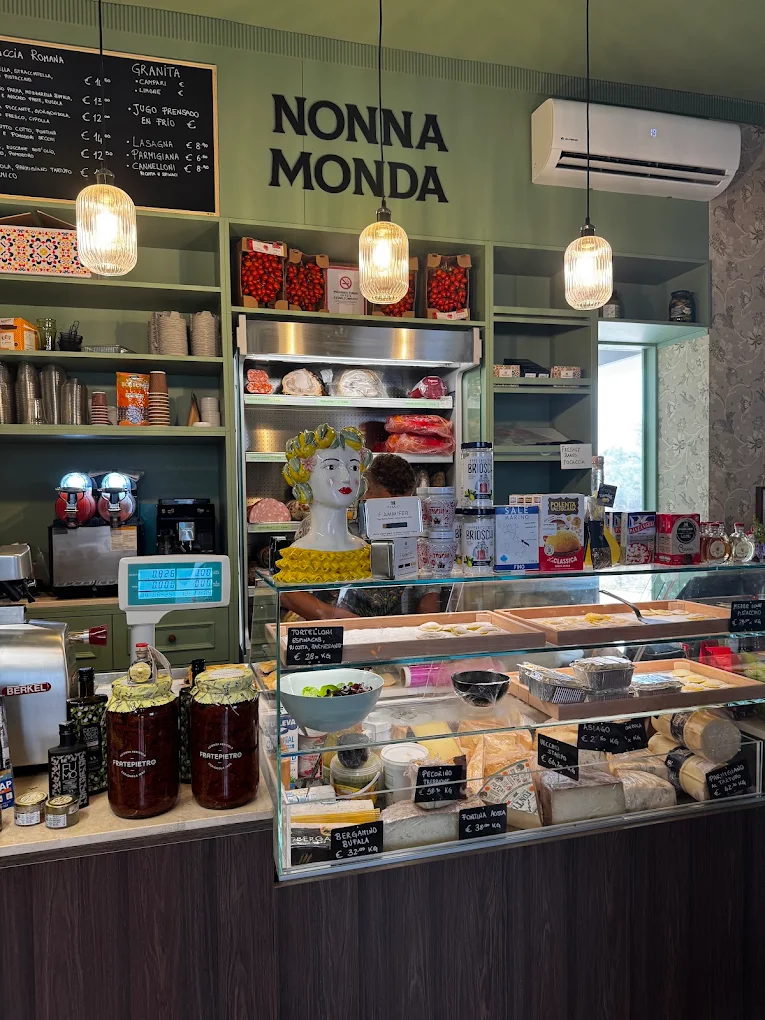 The deli counter: cheeses and cured meats under glass, a red Berkel slicer, jars of preserves and NONNA MONDA lettering on the sage wall above.