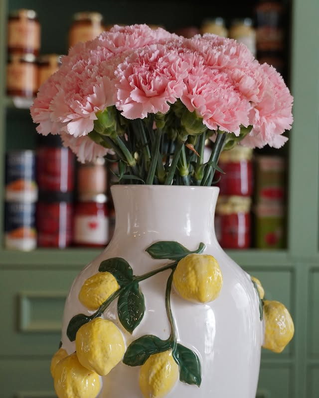 Pink carnations in a white ceramic vase moulded with lemons, in front of shelves of preserves.