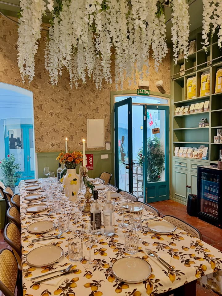 A long table laid with a lemon-print cloth, candles and flowers under hanging white blossom, with the shop shelves alongside.